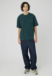 T-shirt oversize vert foncé à col rond et manches courtes, accompagné d'un jean en denim bleu ample et de baskets grises.