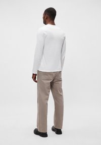 Balta ilga rankovė marškiniai su apvaliu iškirpimu, derinama su smėlio spalvos plačiomis kelnėmis ir juodais batais, matoma iš nugaros.