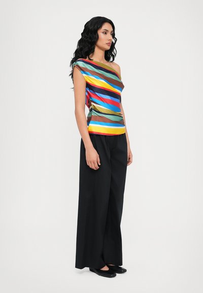 Multicolore one-shoulder top met horizontale strepen in verschillende tinten, gecombineerd met zwarte wijde broek en vlakke zwarte schoenen.