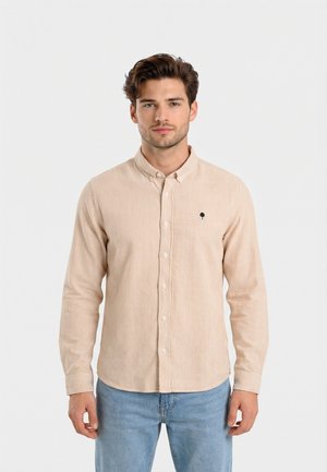 Joven con cabello castaño que lleva una camisa de botones beige a rayas y jeans azul claro, de pie contra un fondo blanco liso.