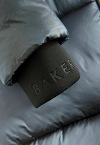 Chaqueta acolchada con un exterior suave de color gris, que presenta un puño elástico negro con la inscripción "BAKER" en relieve. Textura suave y diseño aislante.