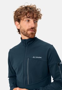 Donkerblauwe Vaude-jack met een ritssluiting aan de voorkant, hoge kraag en een ruitpatroon op het onderste gedeelte, met reflecterende accenten.