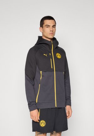 Sort og grå lynlåshoodie med høj krave, gule accenter, logo på brystet, og matchende sorte shorts med sidelogo.