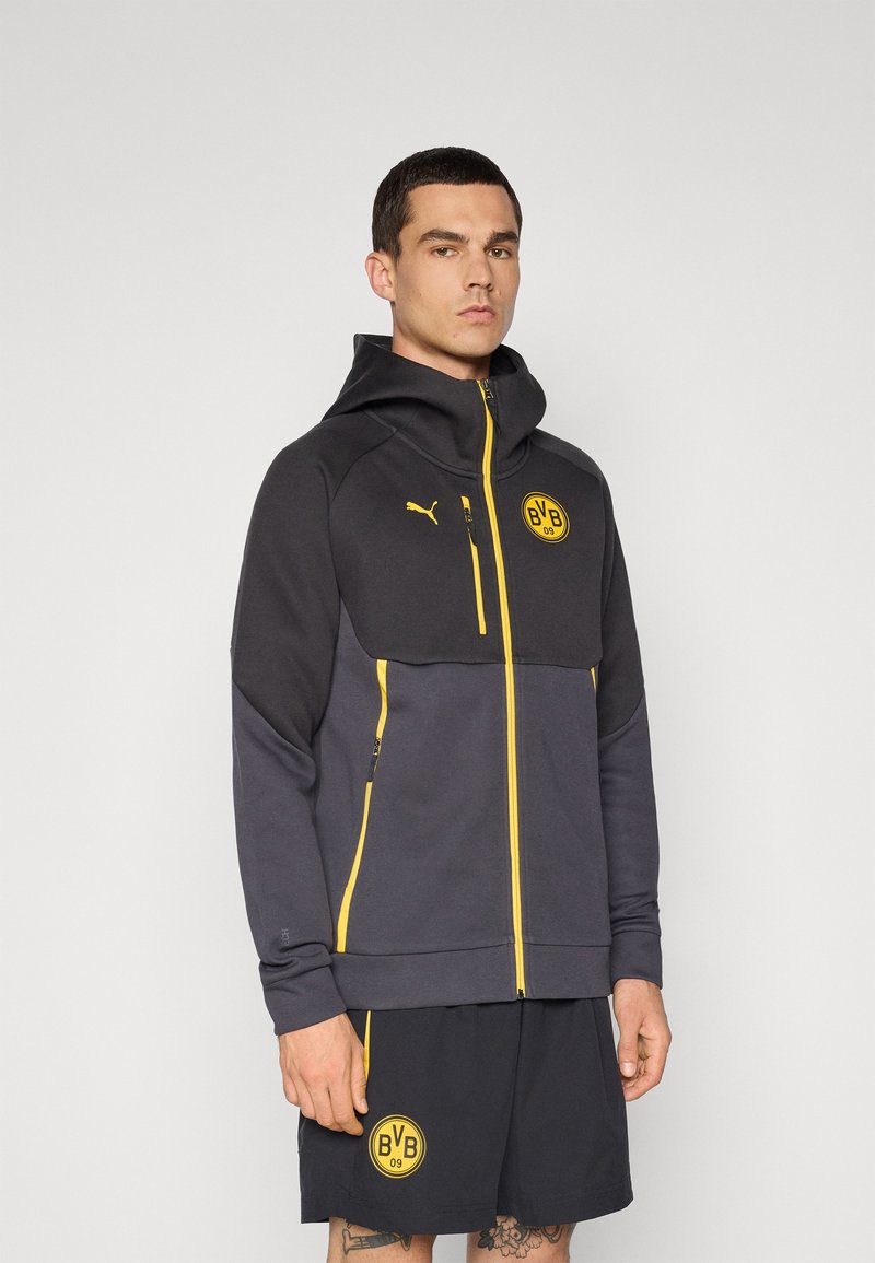 Sort og grå lynlåshoodie med høj krave, gule accenter, logo på brystet, og matchende sorte shorts med sidelogo.