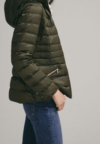 Doudoune vert olive à design matelassé, poches zippées et capuche. Le tissu a une finition lisse et brillante.