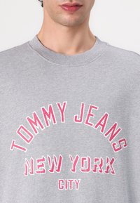 Gray sweatshirt with ribbed collar, featuring bold pink and white text: “TOMMY JEANS NEW YORK CITY” in a curved design. Soft texture.