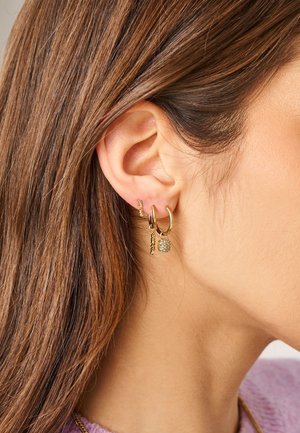 Rechteroor met drie gouden oorbellen: een kleine ring met een getextureerde hanger, een middelgrote ring met een fonkelende kubus, en een kleine ring met woorddetail.