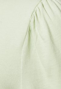 Tessuto a maglia verde chiaro con dettaglio arricciato sulla spalla, che mette in evidenza una texture liscia e una leggera lucentezza.