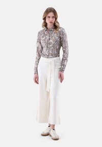 Blusa a maniche lunghe a fantasia in marrone e crema, abbinata a pantaloni bianchi a vita alta con una cintura a cravatta e scarpe beige.