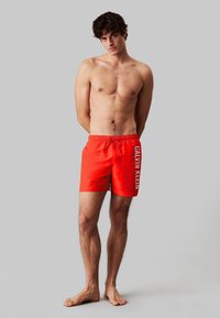 Bañador rojo con cintura con cordón, que presenta un logo lateral en blanco. El material parece suave y ligero.