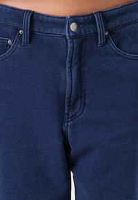 Mörkblå denimjeans med knappstängning, framfickor och synlig sömnad. Tyget har en slät, något strukturerad yta.
