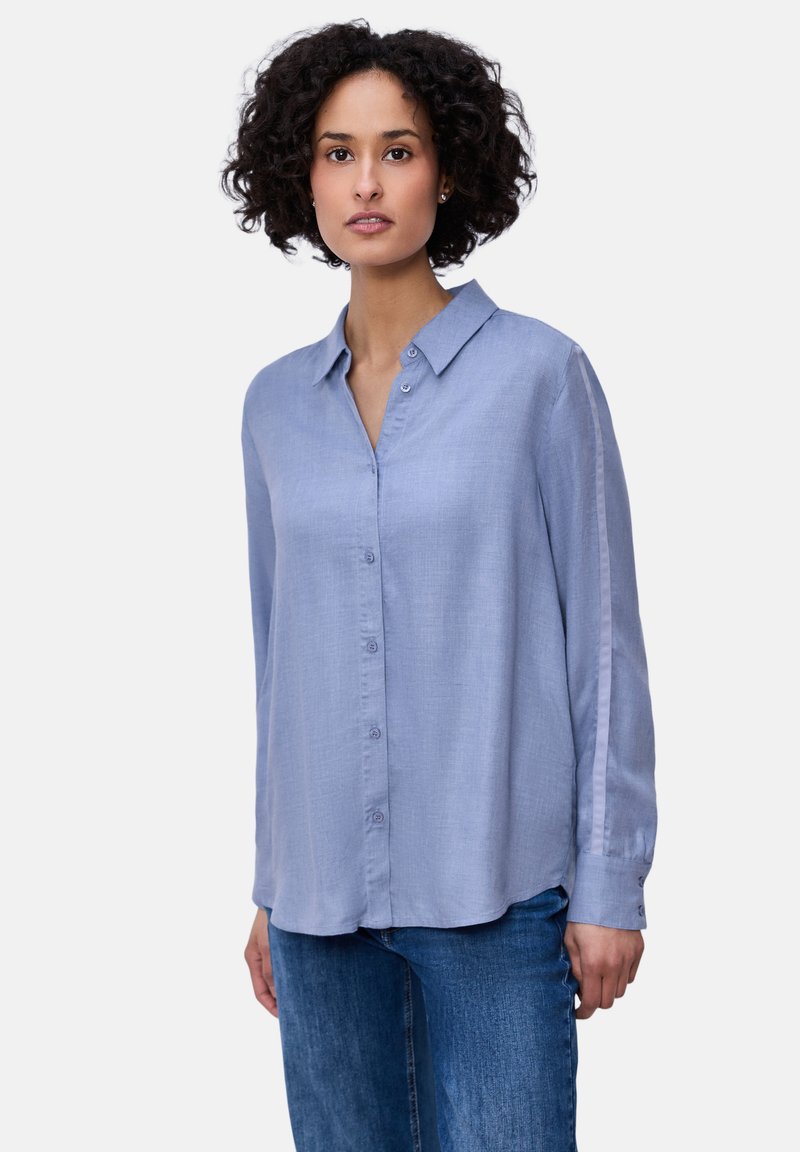 Femme aux cheveux bouclés portant une chemise à manches longues bleu clair boutonnée et un jean bleu, debout devant un fond blanc uni.