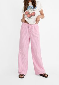 Pantalons larges rayés rose et blanc avec une taille à cordon. Porté avec un t-shirt graphique et des sandales, montrant une tenue décontractée.
