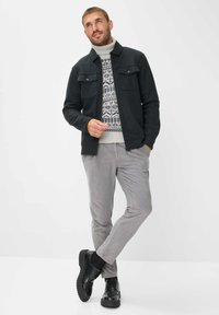 Donkergrijze jas met twee borstzakken, gedragen over een gebreid patroontrui. Het model draagt lichtgrijze corduroy broek en zwarte schoenen.