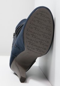 Mörkblå högklackad sko i mjukt tyg, med dragkedja på sidan och ett mönstrat gummisula med varumärkeslogotyp.