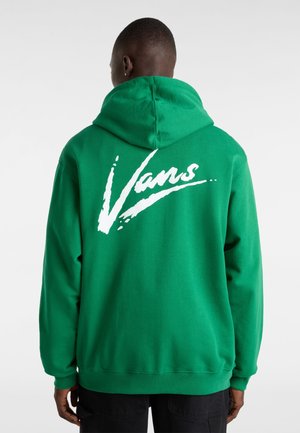 Mand iført en skriggrøn hættetrøje med stort hvidt Vans-logo på ryggen, stående mod en ensartet lys baggrund.