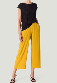 Top corto nero a maniche corte con un design testurizzato, abbinato a pantaloni larghi plissettati giallo brillante. Sandali neri con cinturino completano il look.