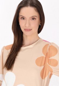 Beige top med store blomster mønstre i orange og hvid, rund halsudskæring og pufærmer med pink accent. Glat stofstruktur.