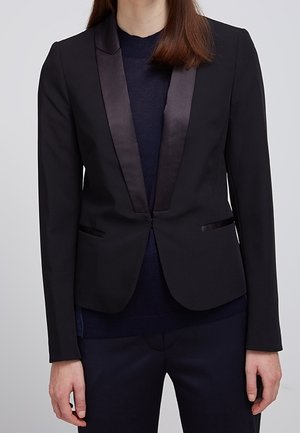 Vrouw draagt een nauwsluitend zwart colbert met satijnen revers over een donkere trui en bijpassende zwarte broek, staande tegen een effen achtergrond.