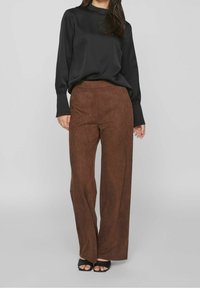 Braune Wildleder-Hosen mit weitem Bein, kombiniert mit einem schwarzen Satinhemd mit langen Ärmeln. Das Hemd hat enganliegende Ärmelbündchen und einen hohen Halsausschnitt.