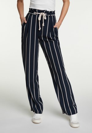 Pantalon bleu marine à rayures verticales blanches, ceinture en corde, poches latérales, porté avec des baskets blanches et un haut blanc rentré.