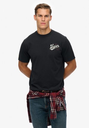 Zwarte katoenen T-shirt met korte mouwen, met een wit en rood logo op de borst. Gecombineerd met blauwe jeans en een rood geruit overhemd om de taille geknoopt.