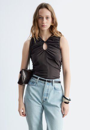 Jeune femme portant un haut noir sans manches froncé, un jean taille haute bleu clair, un sac noir à l'épaule et des bracelets.
