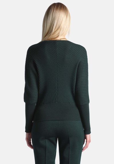Personne blonde vue de dos portant un pull côtelé vert foncé et un pantalon foncé assorti sur un fond uni.