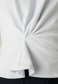 Tissu blanc tordu et rassemblé à la taille, créant des plis et de la texture, sur un pantalon sombre.