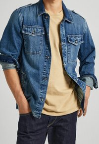 Jeansjacka i mörkblå färg med två bröstfickor, knappstängning och uppvikta ärmar. Bärs över en ljusbeige t-shirt.