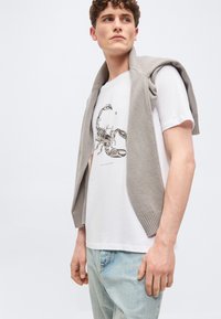 Νεαρός άνδρας με λευκό t-shirt που έχει εκτυπωμένη μαύρη σκορπιό και γκρι πουλόβερ περασμένο στους ώμους του, ανοιχτόχρωμο μπλε τζιν.