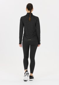 Kvinde med en fletning, der går væk iført en sort prikket langærmet trøje, matchende leggings og sorte træningssko på en hvid baggrund.