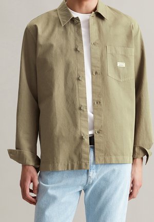 Homme portant une chemise marron clair boutonnée sur un tee-shirt blanc et un jean bleu clair, la poche de la chemise a une petite étiquette blanche.