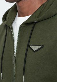 Sudadera verde con cremallera y textura suave, cordones negros, cremallera plateada, y un parche de logo de marca en forma de triángulo en el lado izquierdo del pecho.