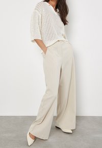 Pletený krémově zbarvený svetr s krátkými rukávy a otevřeným vzorem; kombinovaný se světle béžovými širokými kalhotami s proužky a lodičkami s špičatou špičkou.