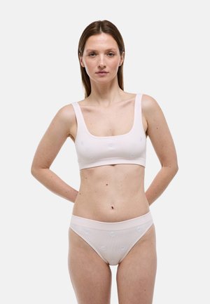 Femme debout, mains dans le dos, portant un soutien-gorge blanc côtelé et un sous-vêtement assorti ornés de petites marguerites brodées sur un fond uni.