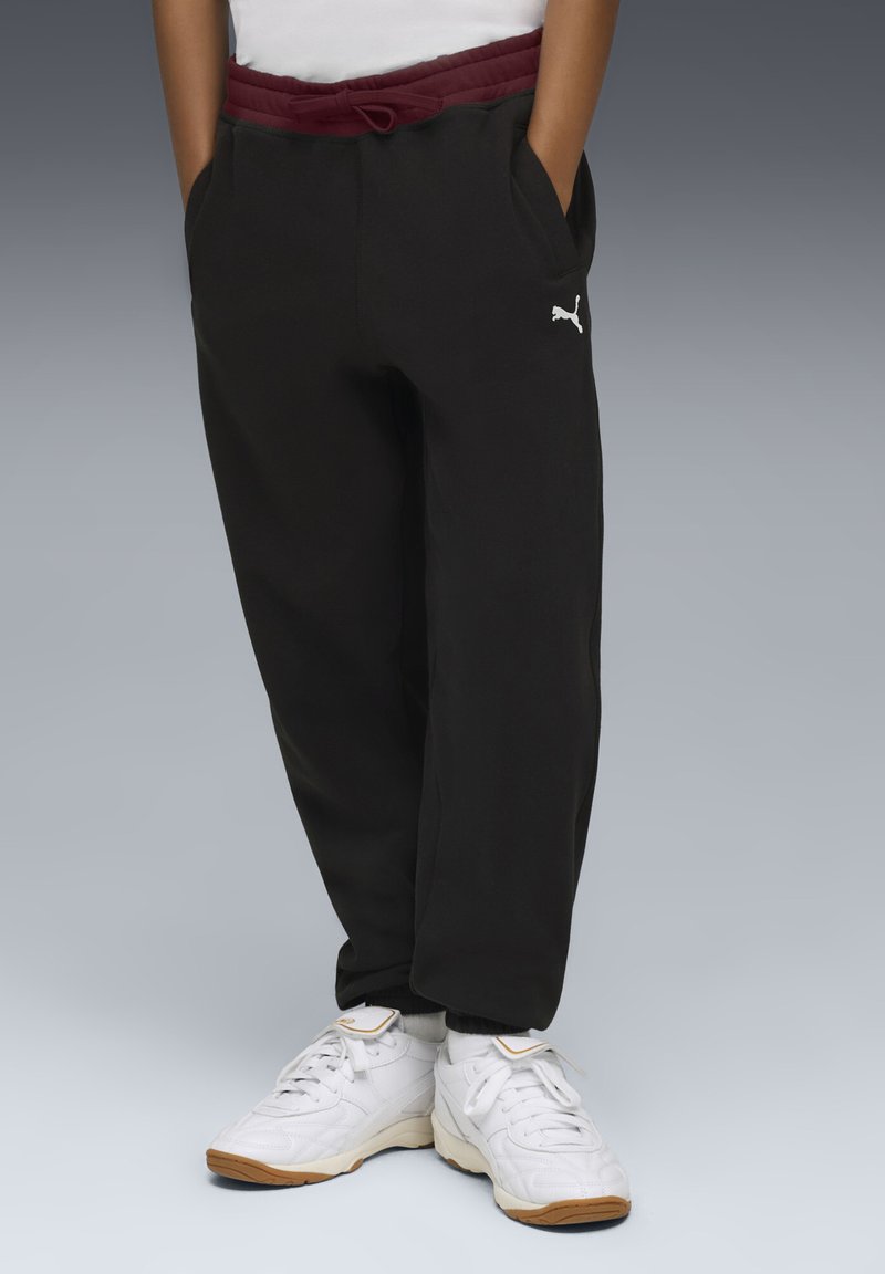 Sorte sweatpants med bordeaux farvet linning, der har lommer og et hvidt logo på venstre side, parret med hvide sportssko.
