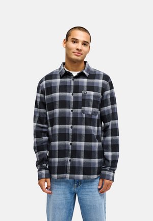 Jeune homme aux cheveux courts portant une chemise à carreaux noire et grise boutonnée et un jean bleu clair, debout devant un fond uni.
