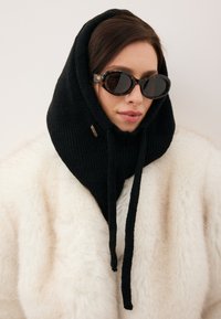 Czarny prążkowany kaptur szalik z troczkami, noszony na białym płaszczu z sztucznego futra; w połączeniu z ciemnymi okularami przeciwsłonecznymi w oprawkach przypominających skorupę żółwia.