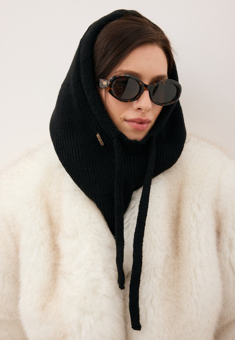 Czarny prążkowany kaptur szalik z troczkami, noszony na białym płaszczu z sztucznego futra; w połączeniu z ciemnymi okularami przeciwsłonecznymi w oprawkach przypominających skorupę żółwia.