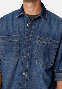 Donkerblauwe denim overhemd met twee borstzakken, knoopsluiting met metalen onderdelen en een klassieke kraag met een gestructureerde afwerking.