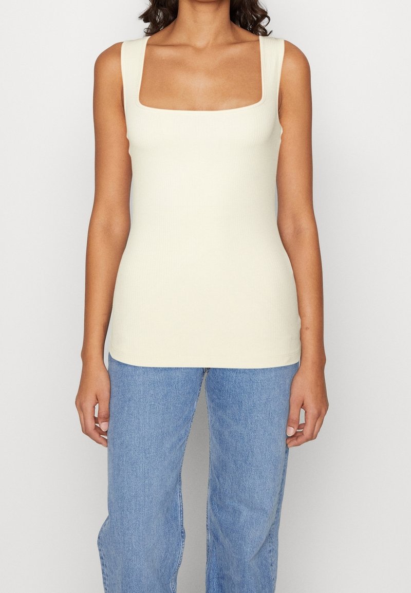 Femme portant un débardeur côtelé crème sans manches avec un col carré et un jean taille haute bleu clair sur un fond uni.