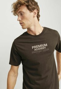 Camiseta marrón oscuro de algodón suave, con una impresión gráfica de color claro que dice "ACADEMIA PREMIUM" y "CONCEPTO INTERNACIONAL."