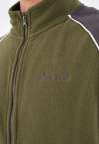 Giacca in pile verde oliva con colletto alto, chiusura con zip frontale e accenti sulle spalle grigi a contrasto. Presenta il logo ricamato "pier one".