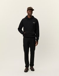 Czarna bluza z kapturem ze sznurkiem i przednią kieszenią, zestawiona z czarnymi spodniami cargo z bocznymi kieszeniami. Brązowe buty o teksturowanej powierzchni i czapka w kratę.