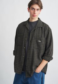 Camisa xadrez de flanela em verde escuro e preto com padrão de grelha branca. Apresenta colarinho, bolso e botões. Usada por cima de uma t-shirt escura.