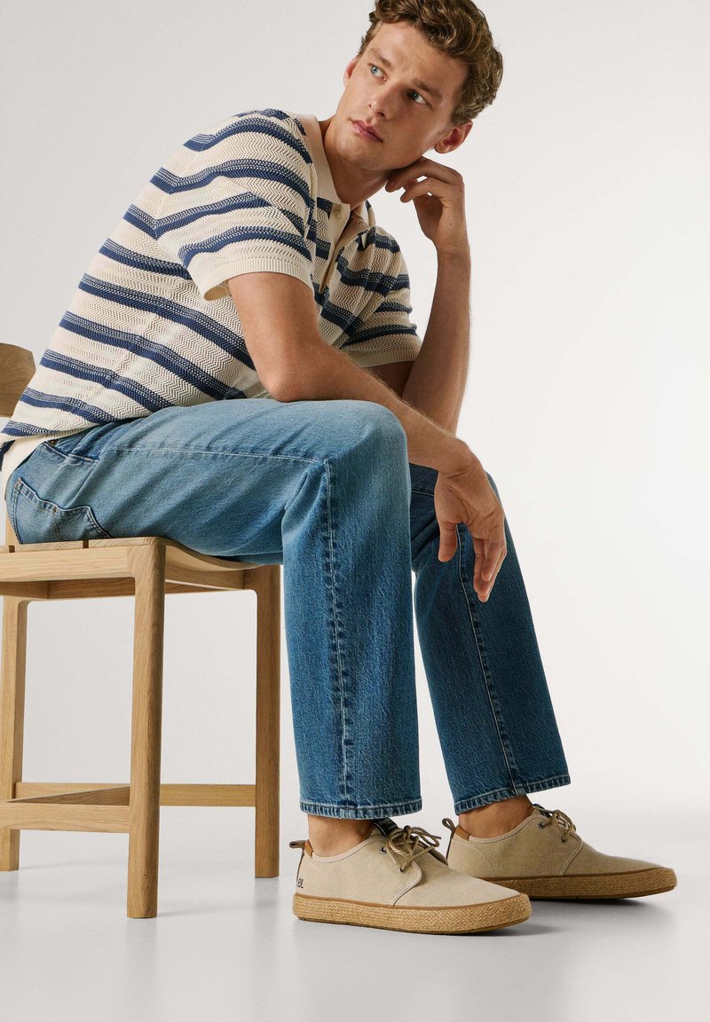 Jeune homme assis sur une chaise en bois, portant un pull rayé tricoté, un jean bleu et des chaussures beiges à lacets, regardant pensivement sur le côté.