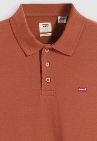 Cămașă polo Levi's de culoare ruginită, cu guler, trei butoane, țesătură texturată și un mic logo roșu pe partea stângă.