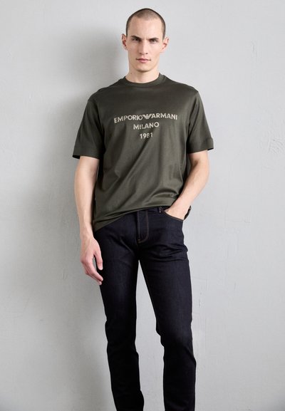 T-shirt en coton vert olive avec logo imprimé "Emporio Armani MILANO 1981", associé à un jean en denim foncé avec des coutures orange.