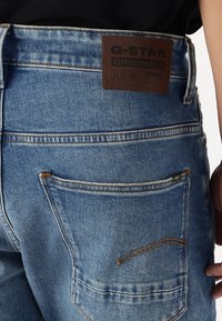 Vaqueros de mezclilla azul con un parche de cuero etiquetado "G-STAR ORIGINALS RAW DENIM". Presenta costuras en el bolsillo trasero y un ligero desgaste.